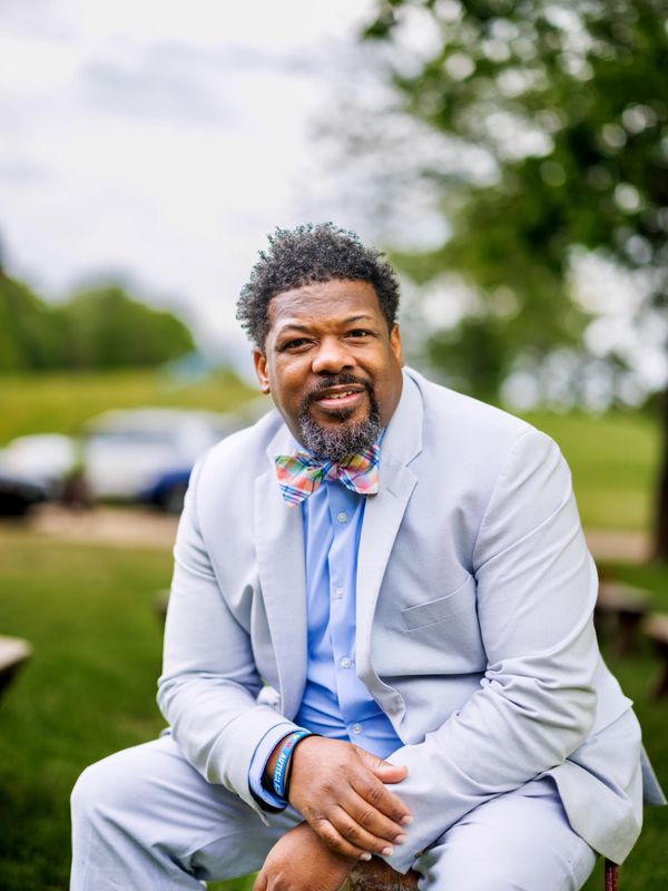 Man in light gray suit with colorful bow tie sitting outdoors.