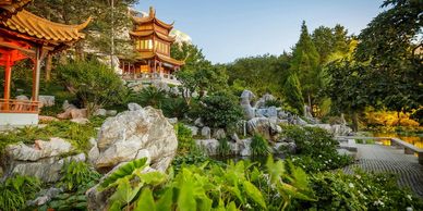 A serene Chinese garden with a pagoda, lush greenery, rocks, and a clear blue sky.