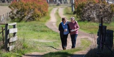 Two women walking arm in arm on a rural dirt path surrounded by greenery.