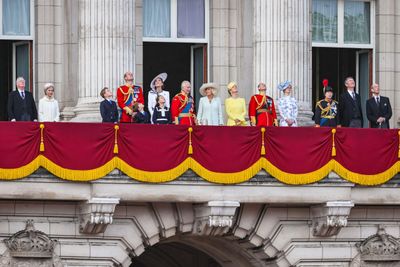 A royal family stands on a balcony adorned with red and gold drapes, looking upwards.