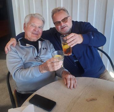 Two older men smiling and toasting with drinks at a table.