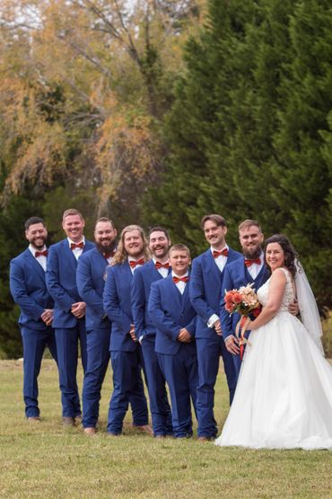 Bride and groom with groomsmen in blue suits and red bow ties outdoors.