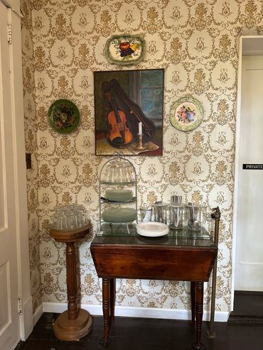 Vintage room corner with patterned wallpaper, a violin painting, decorative plates, and glassware.