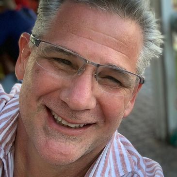 Picture of owner smiling. Male, salt and pepper hair, wire frame glasses and a red stripe shirt