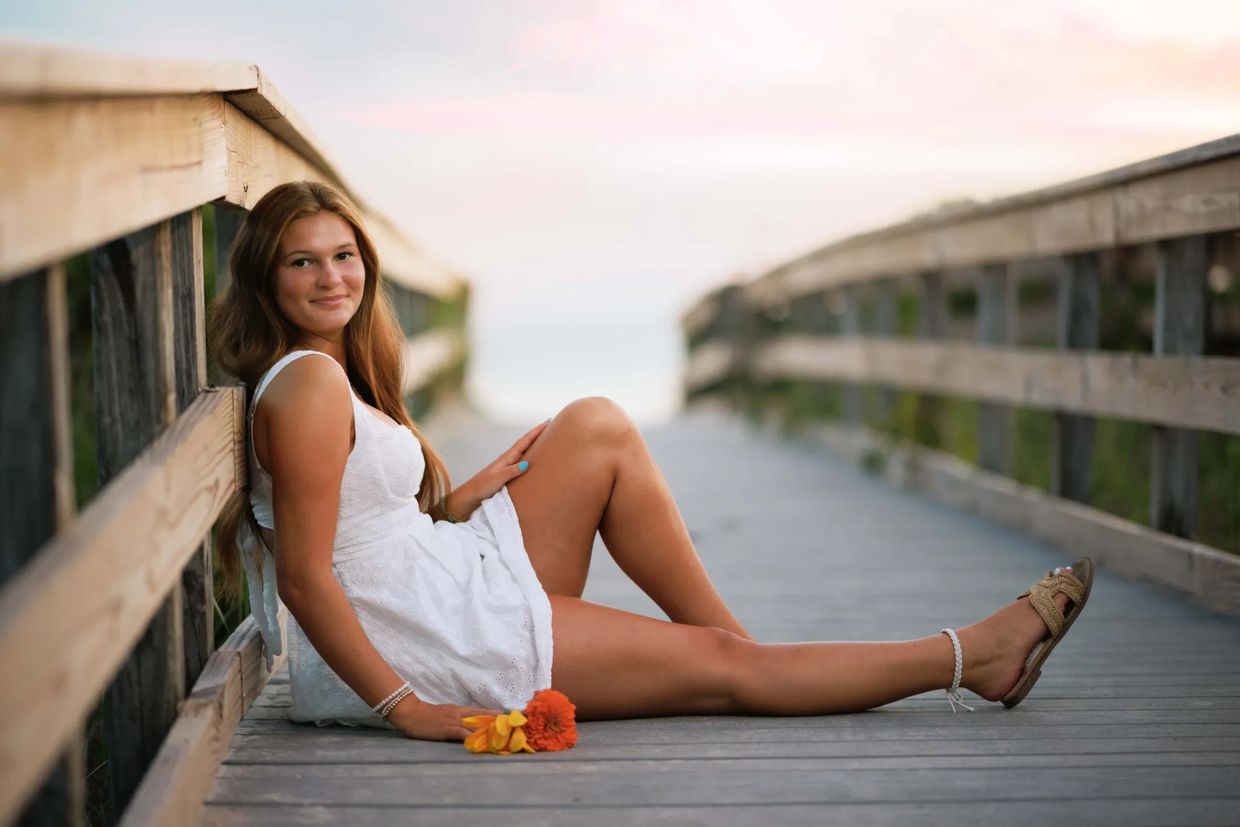 Young woman in white dress sitting on wooden boardwalk with flowers.