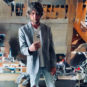 Man taking a mirror selfie wearing a gray blazer and ripped jeans.