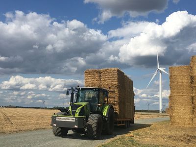 Tractor carrying hay bales near wind turbines under cloudy sky.