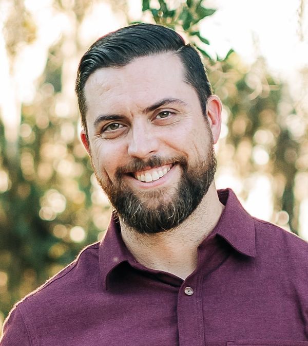 Smiling man with beard in a maroon shirt outdoors.