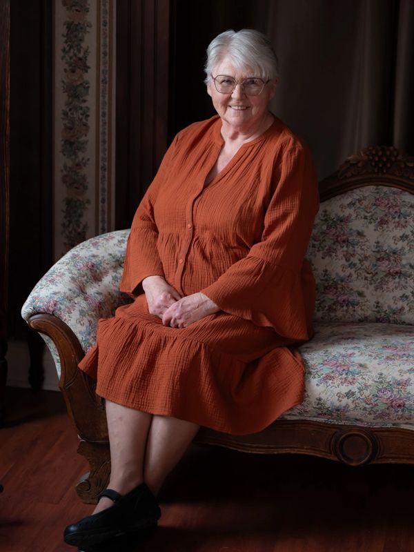 Smiling elderly woman in orange dress sitting on floral vintage sofa.