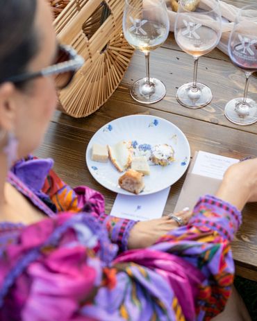 Person in colorful attire enjoying a cheese tasting with wine glasses on a wooden table.
