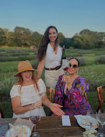 Three women enjoying wine and laughter outdoors at a rustic table.