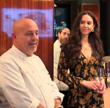 A chef speaks while a woman in a floral dress listens, holding a glass of wine.