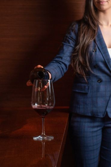 A woman in a blue suit pours red wine into a glass on a wooden table.