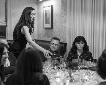 A woman pours wine at a dinner party surrounded by friends in a cozy setting.
