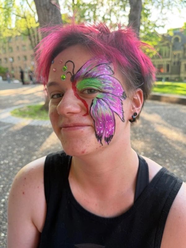 Person with pink hair and butterfly face paint smiling outdoors.