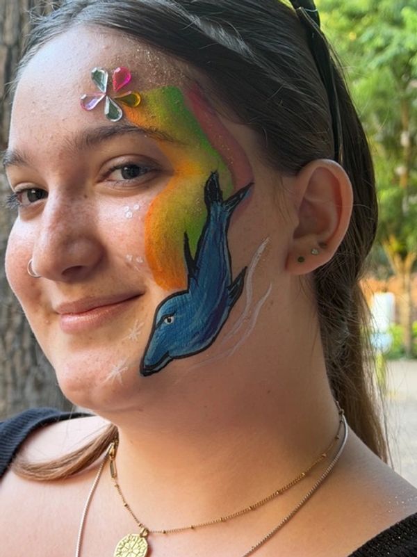 Smiling woman with colorful dolphin face paint and decorative gems on her cheek.