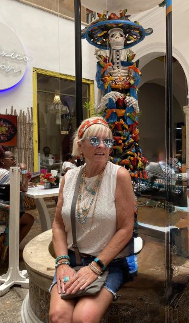 Woman in sunglasses poses beside colorful Day of the Dead sculpture indoors.