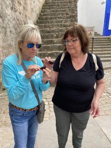 Two women closely examining small objects near stone steps on a cobblestone path.