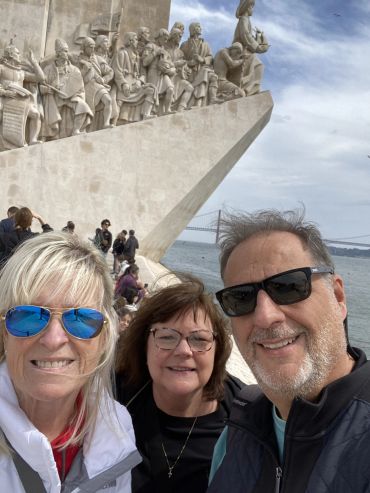 Three people smiling in front of a historic monument by the water on a cloudy day.
