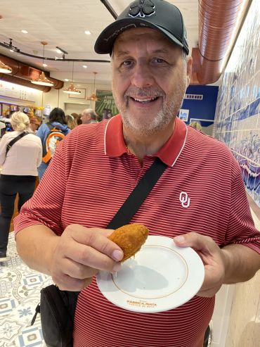 Smiling man holding a fried snack on a plate inside a busy eatery.
