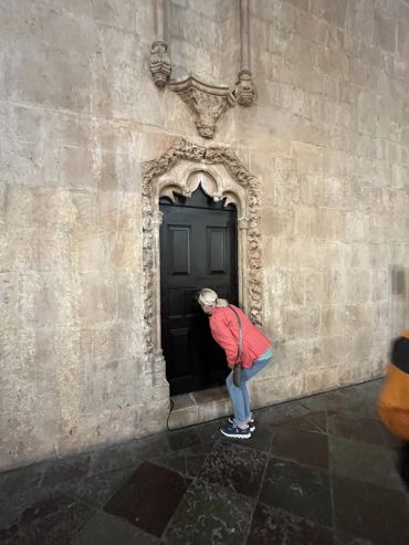 A woman peeks into a small, ornately framed black door in a stone wall.