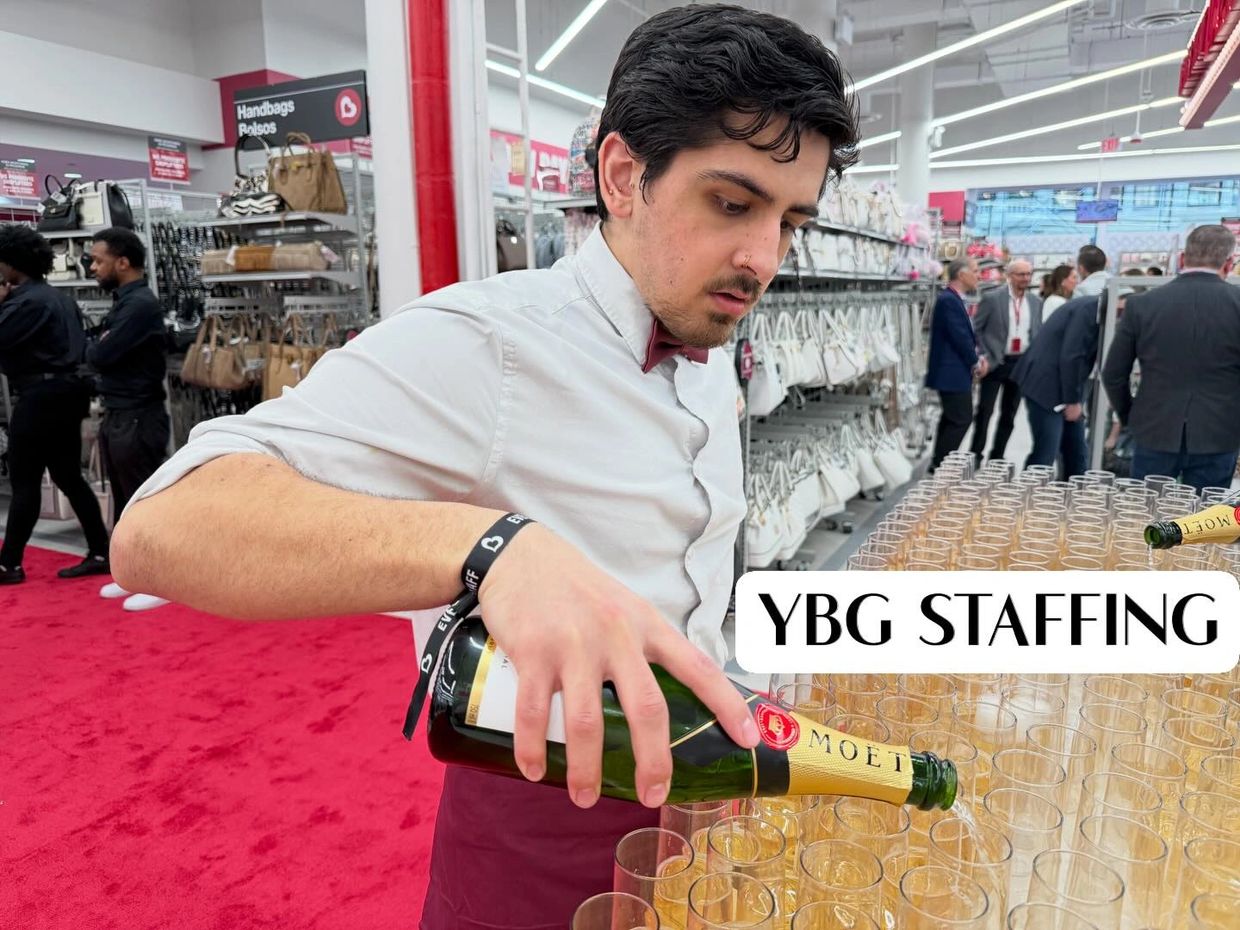 A staff member pours champagne at a busy event with handbags on display.