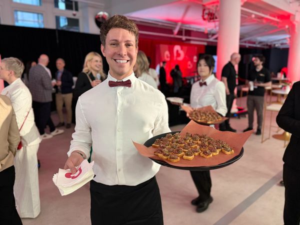 Smiling waiter serving appetizers at a lively event with guests in the background.