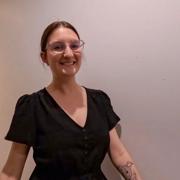 Smiling woman with glasses and a black dress showing a dog tattoo on her forearm.