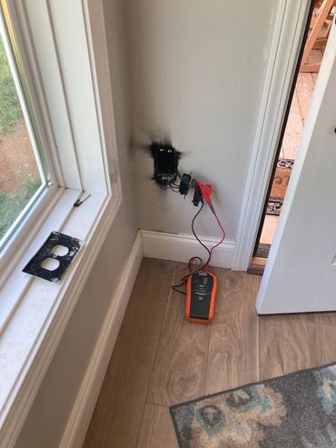 Burnt electrical outlet with wires exposed and a multimeter on the floor beside it.