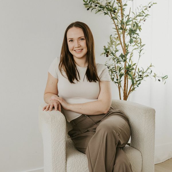 A young woman smiling while seated in a cozy chair beside a tall green plant.