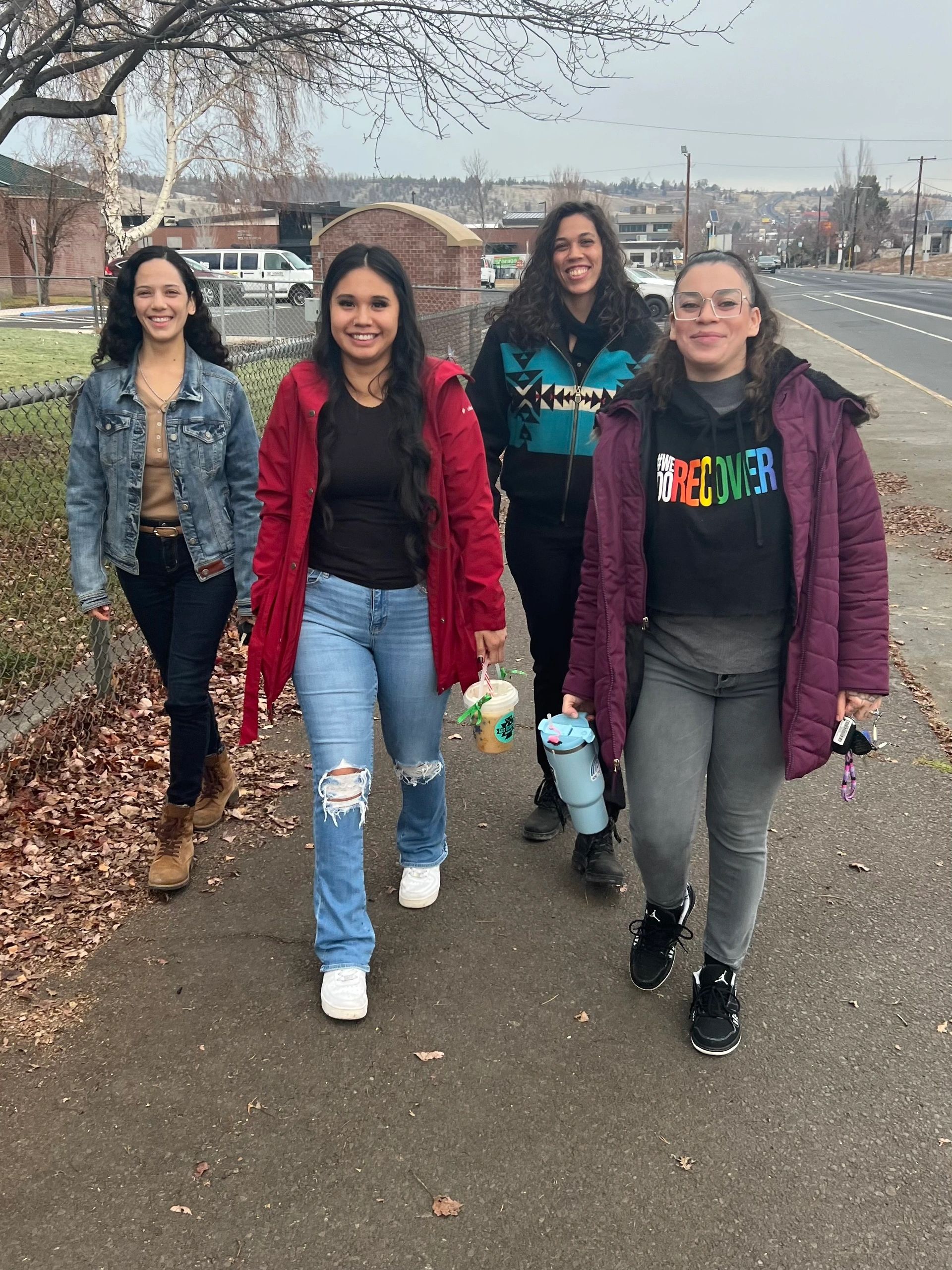 Four women walking happily on a sidewalk in casual winter attire.