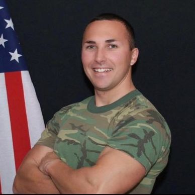 Smiling man in camouflage shirt posing with arms crossed beside American flag.