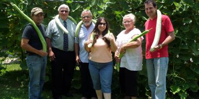 Six people stand together holding long green vegetables in a garden.