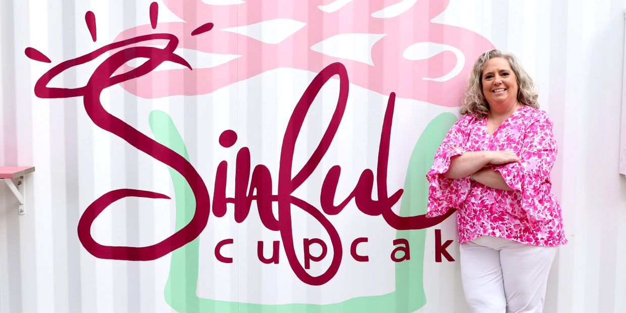 A woman stands smiling in front of a colorful 'Sinful Cupcak' sign.