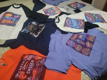 Colorful t-shirts featuring neon-style bar and grill designs laid out on a bed.