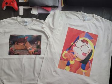 Two white t-shirts with colorful artistic prints laid out on a dark surface.