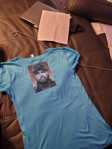 Blue T-shirt with a stylized portrait of a man wearing a cap on a leather couch.
