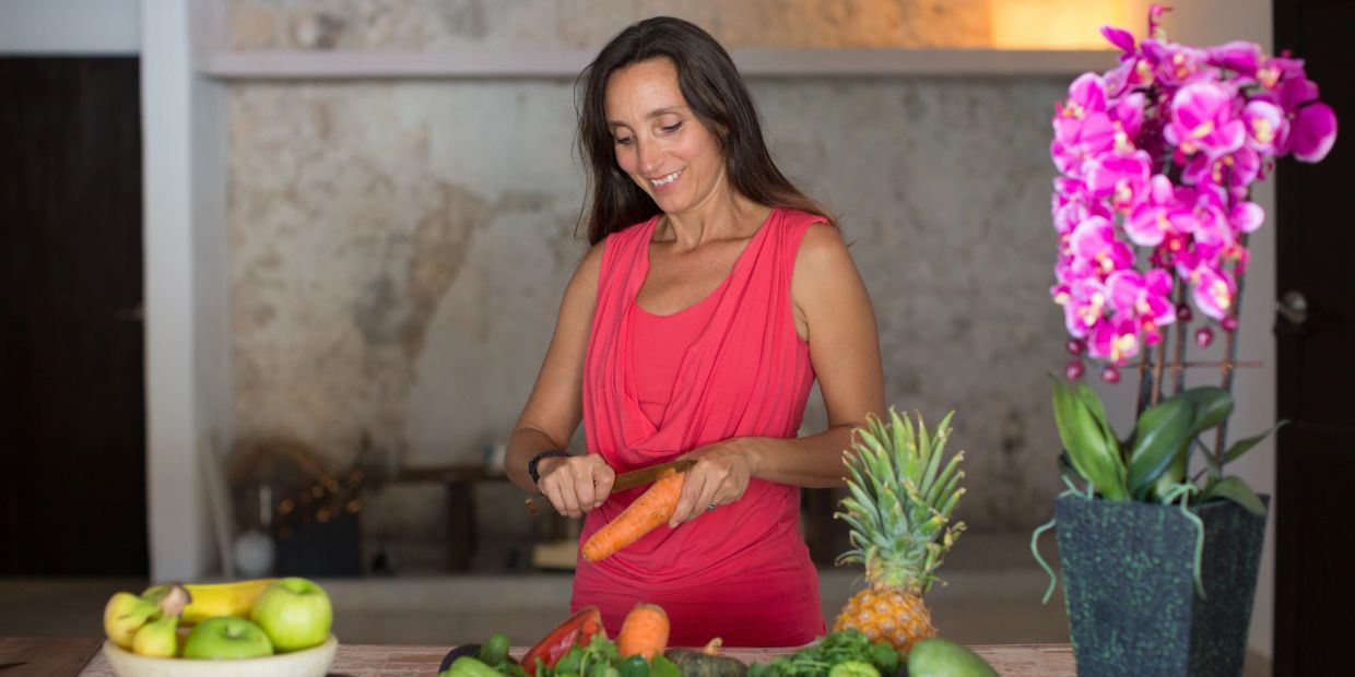 Woman peeling carrot in kitchen with fresh fruits and flowers.