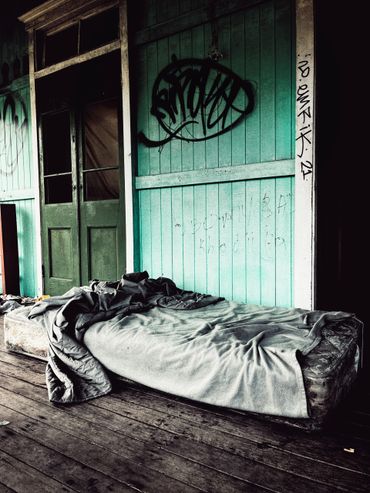 Worn mattress with rumpled bedding on wooden floor beside green wall with graffiti.