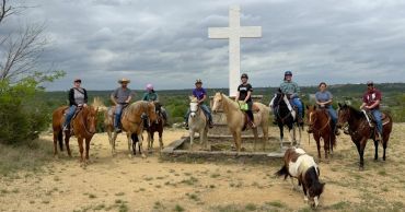 Group of people on horses near a large white cross in a rural area.