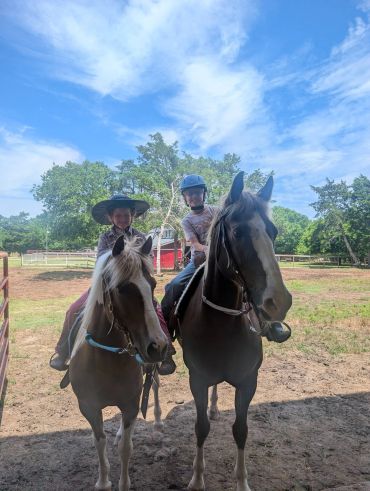 Two children on horseback under a bright blue sky.