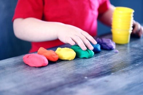 Child playing with rainbow-colored play doh balls on a wooden table.