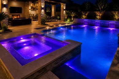 Luxurious backyard pool and hot tub illuminated with vibrant blue and purple lights at night.