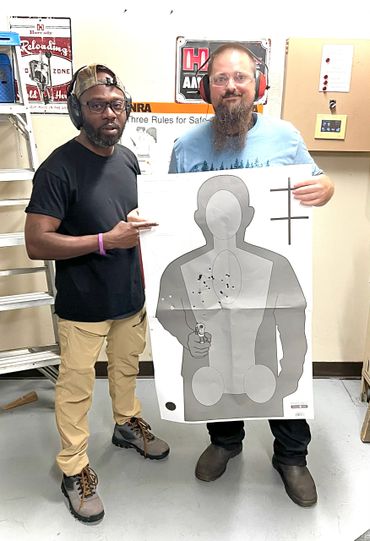 Two men at a shooting range holding a bullet-riddled target silhouette.