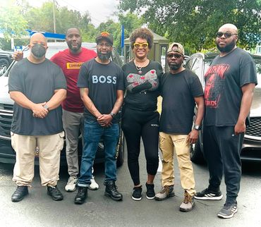 Group of six people standing outdoors, posing in casual clothing with smiles and neutral expressions.