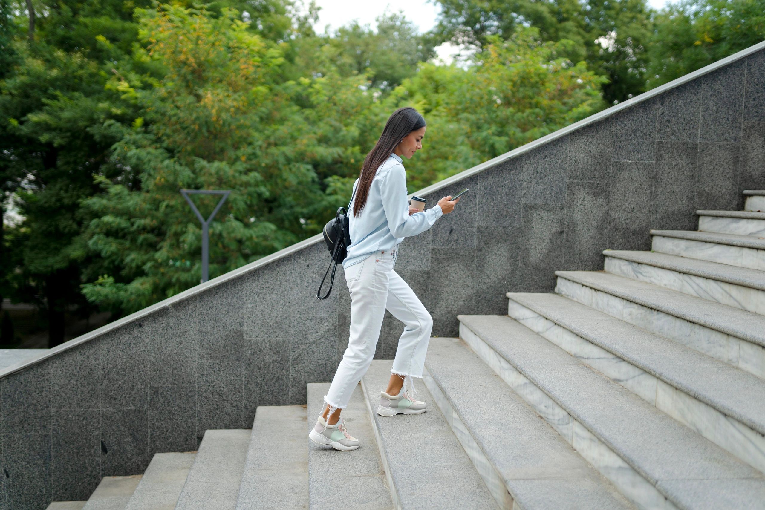 A woman walking up the stairs, wearing white jeans, sneakers, carrying a backpack/purse.