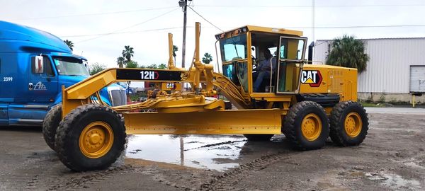 Yellow CAT 12H motor grader parked on muddy ground near a blue truck.