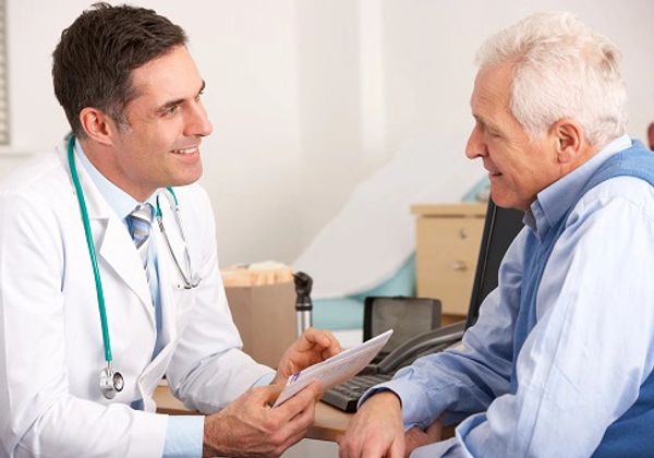 A smiling male doctor in a white coat talks with an elderly male patient during a consultation.
