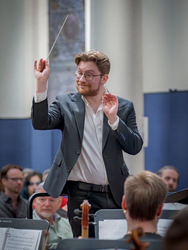 Smiling man in glasses and navy suit conducts an orchestra..