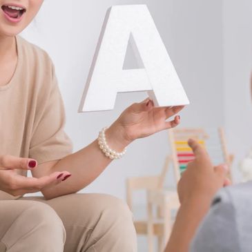 Woman showing decorative block letter A, wearing pearl bracelet, red nail polish to a boy in grey.
